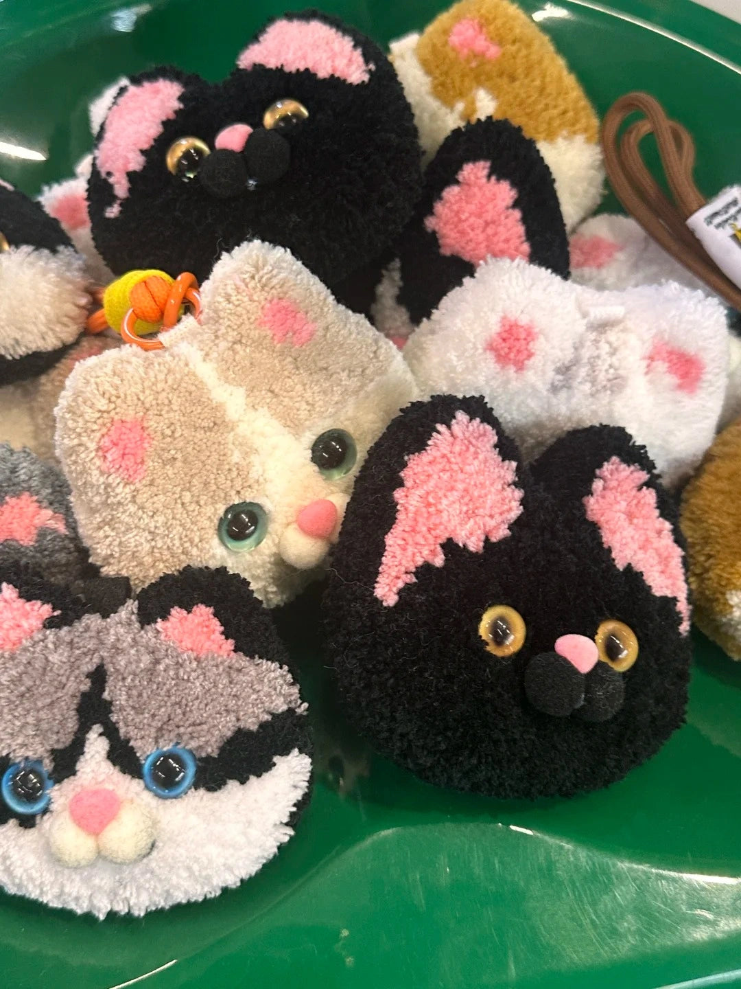 A collection of handmade tufted cat-face plush charms in various colors, featuring soft fluffy textures and round shapes displayed together.