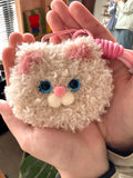 Close-up of a pink fluffy tufted cat-face charm held in hand, showing button-like facial features and soft texture.