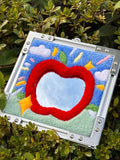 Handmade tufted apple mirror wall art placed outdoors against green leaves, highlighting the bright red apple shape and textured tufted surface.