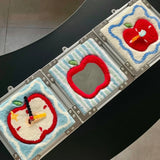 Several handmade tufted apple mirror wall art pieces arranged on a tabletop, showcasing different apple patterns and color combinations.