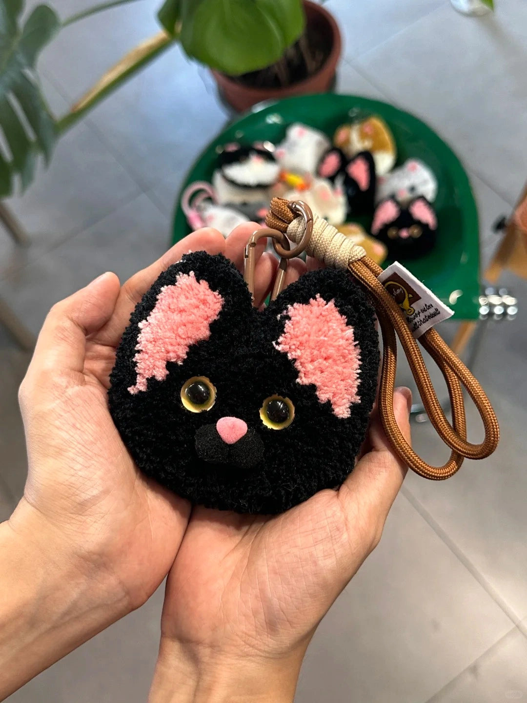 Hand holding a black tufted cat-face charm with pink ears, fluffy texture, and embroidered facial details.