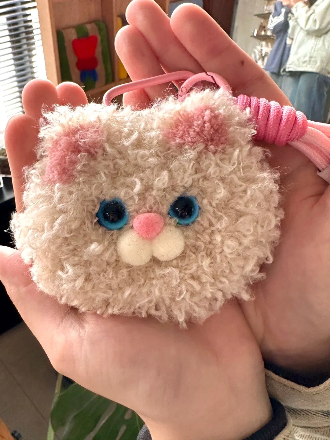 Close-up of a pink fluffy tufted cat-face charm held in hand, showing button-like facial features and soft texture.